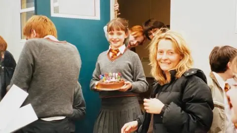 Flick Miles A young girl dressed in a Hogwarts school uniform is smiling holding a birthday cake. She is surrounded by people, some resembling characters from the Harry Potter movies.