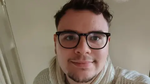 Ethan Appleby A selfie of Ethan Appleby. Ethan has short curly brown hair and a short goatee beard. He wears dark rimmed glasses and a cream pullover. 