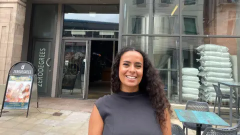 Tia wearing a black sleeveless top is stood outside the Zerodegrees bar in Cardiff with large windows to the right of the bar. She has curly long hair and has a nose ring. A sign can be seen behind her with a table and chairs to her right.