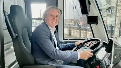 Merseyside Metro Mayor Steve Rotheram sitting behind the wheel of the new glider that will carry passengers around the streets of Liverpool. He is wearing a blue suit and smiling for the camera. 