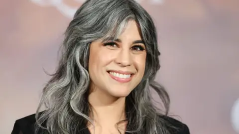 Getty Images A woman with shoulder-length grey-black hair smiles warmly in front of a salmon pink hoarding on a red carpet. 
