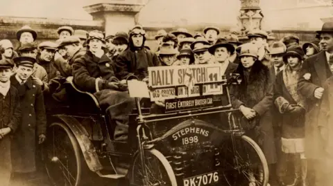 Public domain A crowd of people gathered around a Stephens car. All of them are wearing 1920s outfits