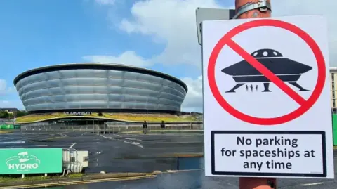 @ASignOfHumour A photo of the Glasgow hydro alongside a sign which says No Parking for spaceships at any time. The joke comes from the spaceship in the sign being a similar shape to the Hydro building