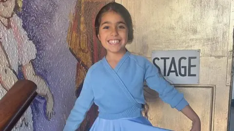 Family photo Image shows Alice Aguiar, wearing a blue cardigan and blue ballet costume. She is standing in front of a sign painted on a door. The sign says 'stage' 