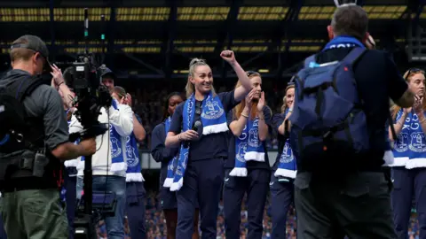 EPA Members of Everton's women's team clap while standing on a podium on the Goodison Park pitch during a ceremony to mark the men's last game at the ground. Howard Kendall Gwladys Street End can be seen in the background.