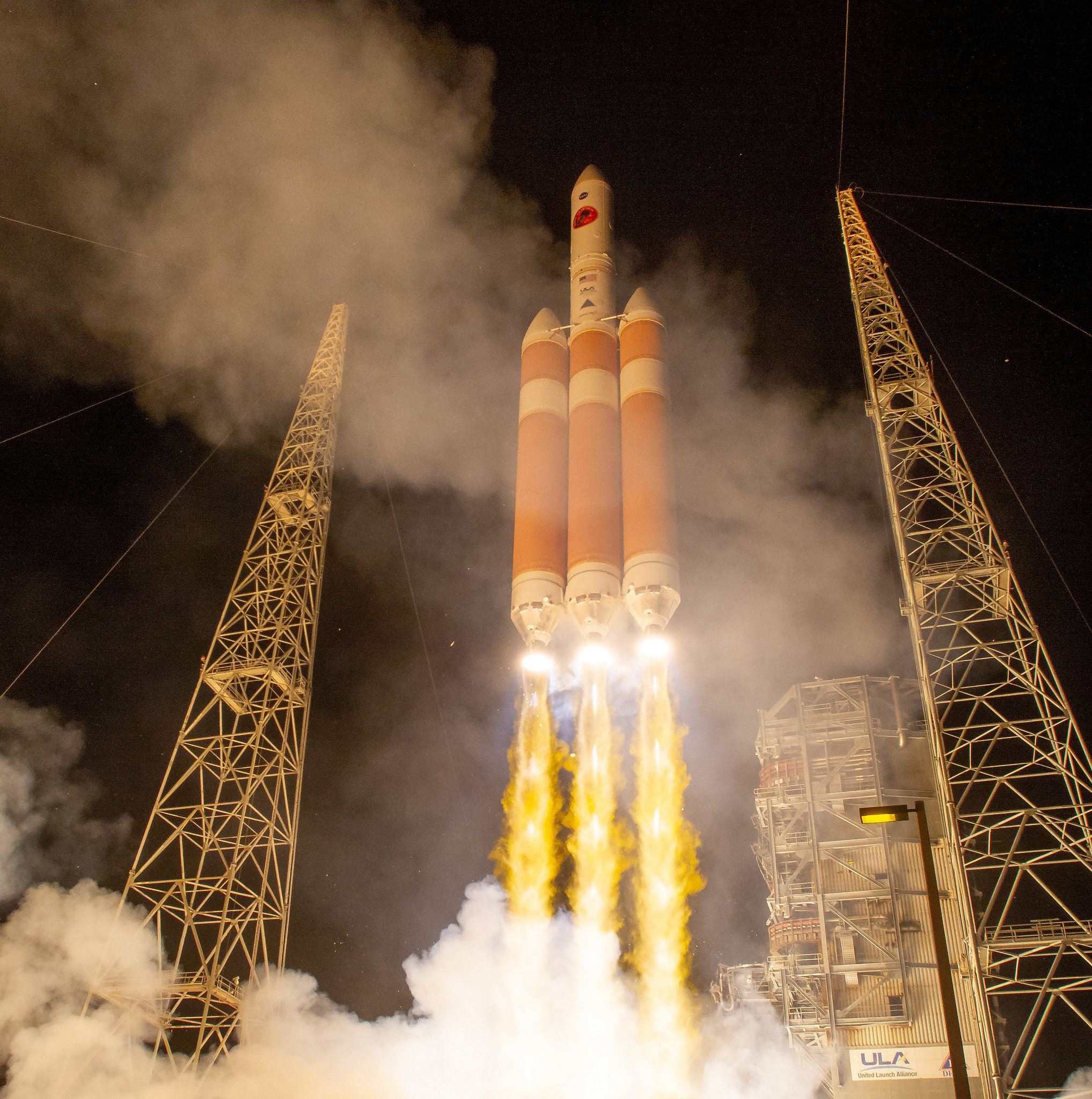 an orange-and-white rocket with two side boosters launches above three plumes of fire between two metal towers