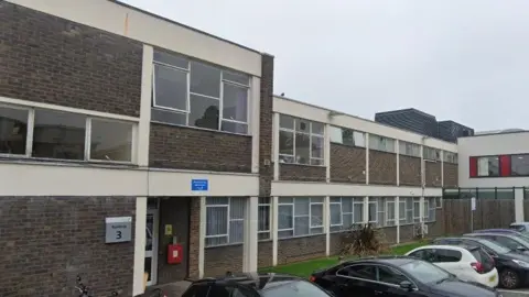 Google A dark brown bricked building goes across the image. The two-storey building has multiple windows and an entrance on the left. Cars are parked outside and a stretch of grass goes alongside the building.