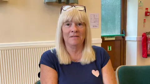 A woman sat on a chair inside a building wearing a navy t-shirt. She has shoulder-length blonde hair and a fringe. She has glasses on resting on the top of her head. She is looking into the camera.