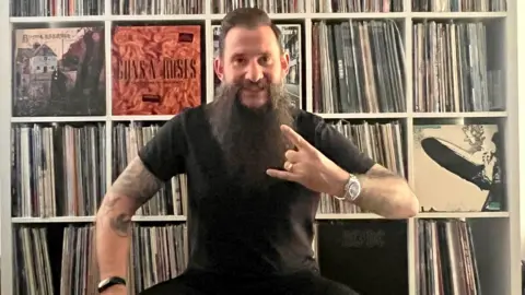 Martin Hughes Martin sitting in front of a shelving unit full of vinyl records. He has a long black beard, arm tattoos and black hair, and is pulling a metal music hand gesture