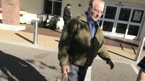 BBC/Mark Horgan A man with thinning hair, glasses, a blue shirt, and a green fleece walks across a car park. He looks down at the ground