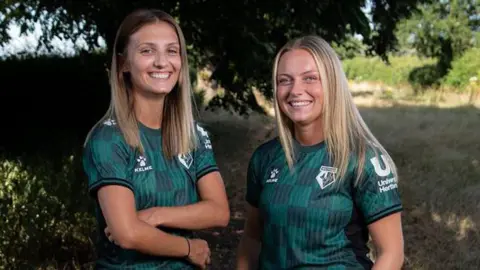 Watford FC Two young women stand in the countryside on a grass track under some large trees. They wear the new Watford away shirt which is a dark forest green with lighter green checks. They smile at the camera and both have long straight fair hair. It is a sunny day.