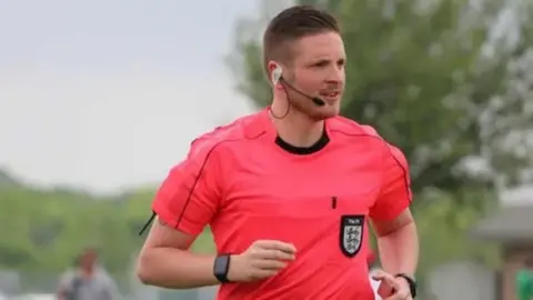 FA A picture of Ryan wearing a pink referee shirt running. He is wearing a microphone on his face and is wearing a watch on both of his wrists.