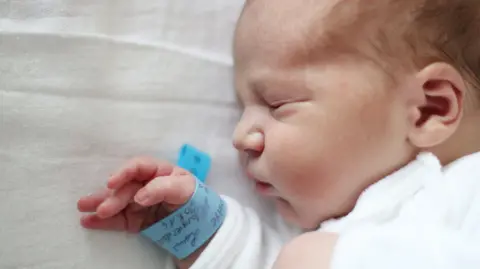 Getty Images A baby boy is sleeping on a white sheet. He is wearing a white sleepsuit and has a blue hospital wristband