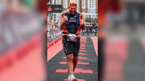James Baker A man celebrates after crossing the finish line at a running event, with people behind a black and red barrier smiling and cheering