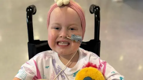 Supplied Eliyah, in hospital, smiling at the camera, in a wheelchair with a tube in her nose