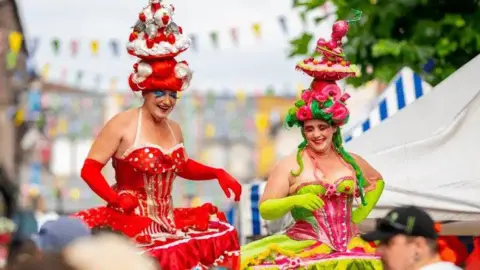 Grum Two people are dressed up on stilts with a market in the background. One has a red wig with a huge pink and red hat in the style of a cake. They are wearing red gloves, blue sparkly eyeshadow and a halter neck red and white puffy dress, The other person has a pink and red cake-style hat, with a green wig. They are wearing a pink and green large dress that billows out.