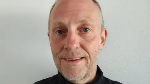 Brian Jenkins A balding man in a black top staring at the camera, he has light stubble that is mostly grey with flecks of black and the top of a black top is visible on his shoulders