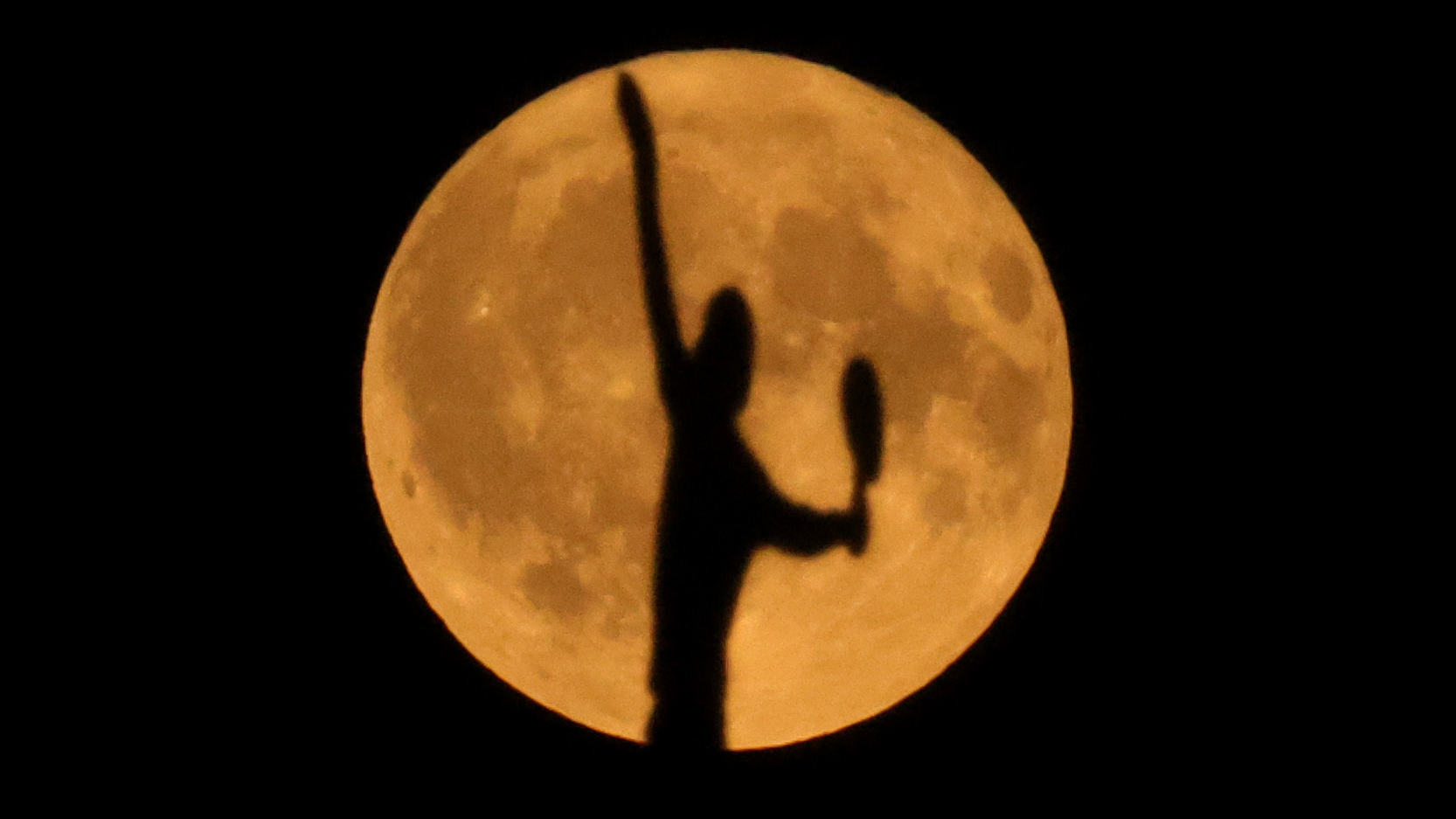 The silhouette of a serving tennis player is seen against the orange disk of a full moon hanging against a dark night sky.