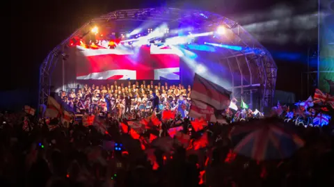 Martin McKay Bedford Proms in Bedford Park, with an orchestra and singer on the stage, the Union Flag behind them, and crowd members, waving their flags. Lights are above the stage.