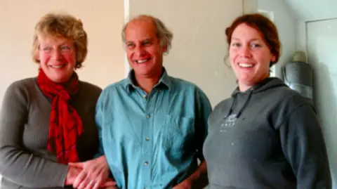Debbie Hemmings A family photo with Ros on the left, Martin in the middle and Debbie on the right. They are standing together with Ros and Martin arm in arm.
Ros is in a grey jumper with a red scarf, Martin is wearing a blue shirt and Debbie is wearing a grey jumper. All are smiling at the camera.