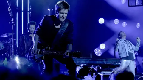 Gavin Elder A guitarist with black clothing on stage with a band and two other members of the band.