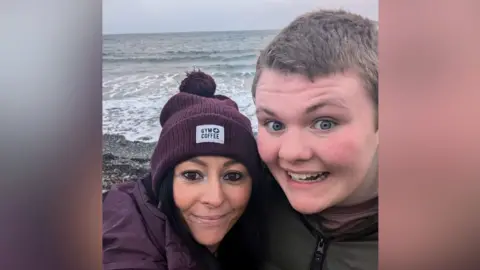 Leanne McCrotter A woman and a boy take a selfie hugging each other. The woman has a purple beanie hat and the words Gym/coffee on it. She has brown eyes, dark brown hair and a purple coat. She's smiling with her mouth closed. The boy is a young teenager but tall and has short brown hair and blue eyes. He's looking at the camera with a wide smile and wide eyes.