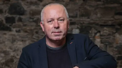 Tim Jarvish/Dervish A bald man in a black blazer and jumper sitting down against a brick wall. He has blue eyes and is half smiling at the camera.
