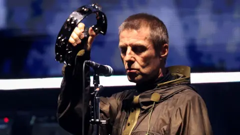 Reuters Liam Gallagher during gig on stage in brown parka holding tambourine up in one hand