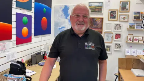 Matt Shepherd/BBC A man with a white beard and glasses is standing in a gallery with a range of different paintings in a variety of styles. Some show bright red and green circles in front of a bright green background. His polo shirt has the logo Market House.