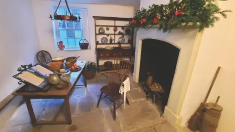 Charles Wesley's House A room inside Charles Wesley's home. There is a kitchen table with some chairs placed next to it. There are some bowls, utensils and a large book on top of the table. There is a fireplace in the room with some wreathes laid on the mantlepiece. In the corner there is a cabinet which is decorated with plates and mugs.
