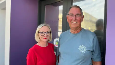 Racheal Day, left, is a petite woman with short bobbed blonde hair, black glasses and red lipstick. She is stood smiling and wearing a red dress. Terry Naftel, right, is a tall man in his 70s/. He has grey hair and glasses and is also stood smiling in a blue t shirt.