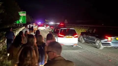 Jodie Halford/BBC People are walking on a pavement, which is on the left. On the right, there is a busy road with traffic. It is dark. The cars have their lights on.
