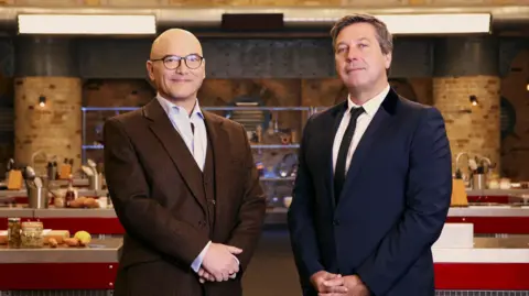 A picture of Gregg Wallace, left, and John Torode, standing in the Masterchef kitchen. Wallace has a shaved head and glasses and is wearing a brown suit with waistcoat, with his hands clasped in front of him. Torode has short dark hair and is wearing a navy suit and tie. Behind them are kitchen cooking stations, with ingredients and kitchen utensils. 