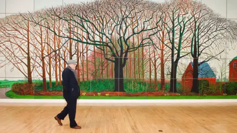 Getty Images Artist David Hockney walking in front of his painting Bigger Trees Near Warter. He is wearing a black jacket, black trousers and brown shoes. He is also wearing a grey flat cap and scarf. The painting behind him is of autumnal trees.
