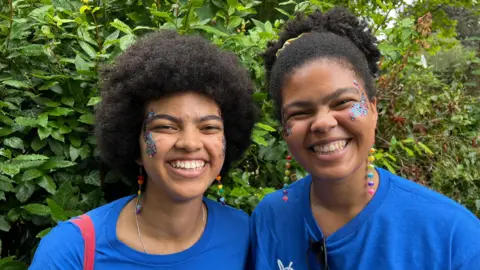 Dolly Kombate (L) and Liz Kombate (R) wearing blue branded Mind t-shirts, matching colourful dangly earrings and blue glitter on their cheekbones. They are standing in front of a bush, happily smiling at the camera.