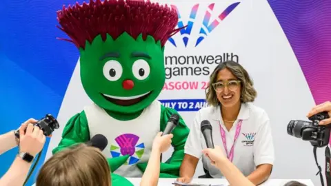 Glasgow 2026 Clyde, an animated thistle mascot with green leaf-like skin and purple spiky hair sitting next to a smily lady wearing glasses and a white polo shirt. Behind them in a banner with the Glasgow 2026 logo on it. In front are people holding cameras and microphones.