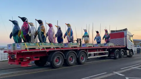Havens Hospices 10 sculptures of penguins, which are all painted different patterns and colours, are standing on the back of a lorry that is travelling down a road.