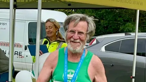 Supplied A joyful looking older man in a green running vest standing outside with cars parked behind him. He has a blue ribbon round his neck, is wearing glasses and has greying hair.