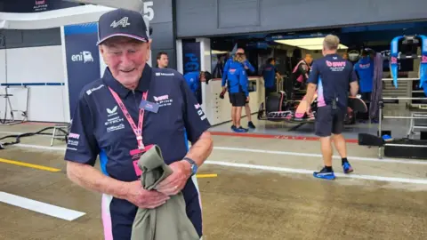 Lucy Wallduck A man stands in front of a Formula 1. He is wearing a navy shirt with pink and white accents and holding a jacket. Team members are visible inside the garage, and the ground appears wet.
