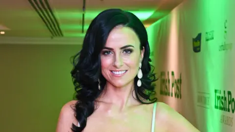 Getty Images A woman with long black curly hair smiles at the camera as she wears a light coloured strappy dress at an event. She has long pearl drop earrings and is wearing makeup.