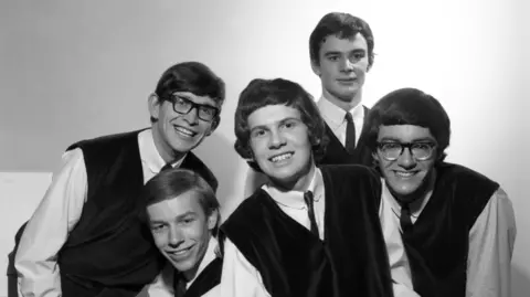 The Zombies when they appeared on Top Of The Pops in 1964. The photo is black and white. The five young men all have dark hair and are wearing loose waistcoats in a dark fabric, along with white collared shirts and skinny ties. Two of the men are wearing glasses. 