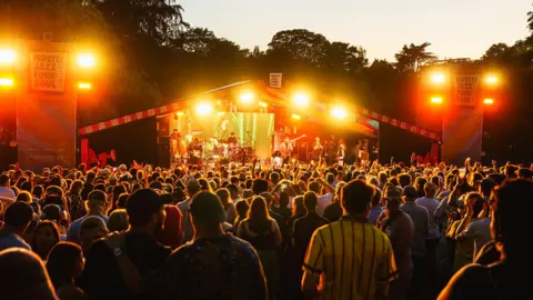 Mostly Jazz, Funk and Soul Festival Crowds gather at a stage with artists playing. Signs on either side of the stage say "Mostly Jazz, Funk and Soul Festival". Orange lights are flashing as the crowd plays