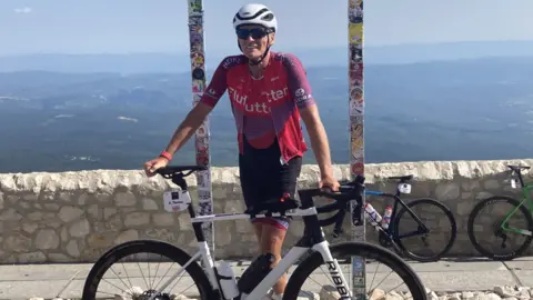 BBC Thomas is stood in full cycling kit leaning on his bike by a low wall with the sea behind him.
