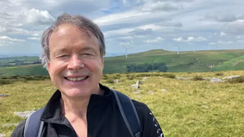 Photograph of Steve Davison from the Rooley Moor Neighbourhood Forum. The picture is taken with the existing Scout Moor wind farm in the background.