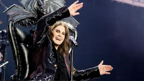 Ross Halfin A man sits on a black throne with a bat motif at the top. He has shoulder-length brown hair and he is wearing black. There is a microphone in front of him.