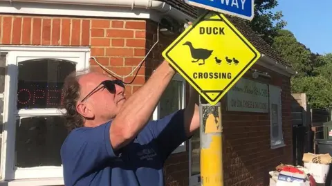 Sarah West A man wearing a blue t-shirt and sunglasses, attaching a yellow diamond-shaped sign to a lamppost using cable ties. The sign says 'duck crossing', with a silhouette image of a mother duck being followed by three ducklings.
