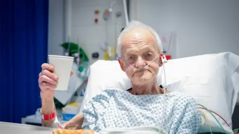 Leeds Teaching Hospitals Trust A man in his late-seventies sits up in a hospital bed and raises a paper cup up to the camera. 