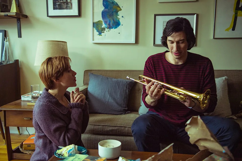 a woman and a man are in a living room, the woman kneels next to a coffee table and looks at the man who sits on the couch and holds a trumpet that he looks at in disbelief