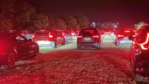 Richard Smith/BBC A long line of cars queueing up on a field. It is dark. The cars' tail-lights are shining red.