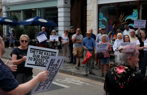 Jonathan Habens Protesters gather in a street. Some are holding placards that say 'they're killing our shop to boost their budget'.